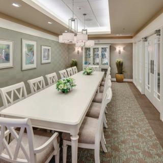 A spacious dining room with a long table surrounded by chairs, decorated with chandeliers and wall art.