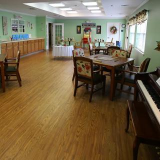 A spacious dining area with wooden floors, multiple tables and chairs, a piano, and large windows.