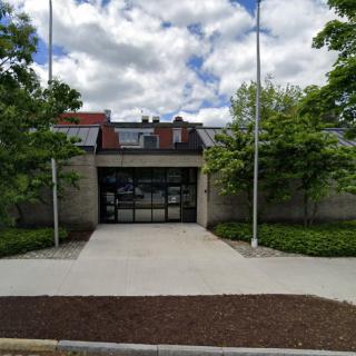 A concrete building entrance with flagpoles, surrounded by trees under a cloudy sky.