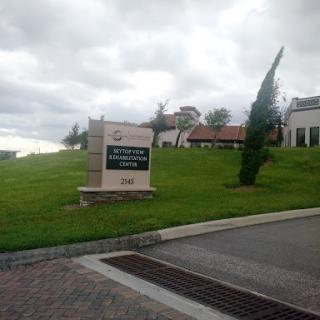 A welcoming sign at the entrance of a building, surrounded by green grass and under a cloudy sky.