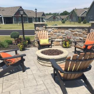 A cozy outdoor patio area with a fire pit surrounded by Adirondack chairs, set against a backdrop of landscaped gardens and charming homes.