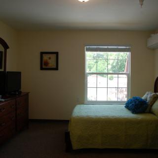 A cozy bedroom featuring a neatly made bed with green bedding and a TV on a wooden dresser, bathed in natural light from the window.
