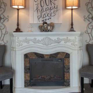 A cozy living room setup featuring an elegant white fireplace, flanked by two chairs and lamps, with a piece of artwork above.