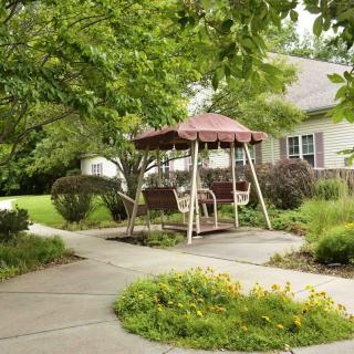 A serene garden setting featuring a covered swing along a winding pathway, surrounded by lush greenery and shrubs near a cozy home.