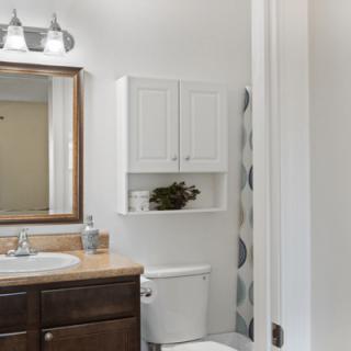A neatly organized bathroom featuring a sink with a mirror, overhead lighting, and cabinetry above the toilet.