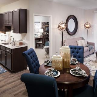 A modern apartment interior featuring a dining area with a round table and blue chairs, adjacent to a kitchen with dark cabinets and a cozy living room.