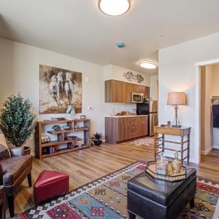 A cozy living area with a connected kitchen, featuring warm wood floors, comfortable seating, and decorative elements like artwork and an indoor plant.