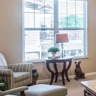 A cozy living room with a large window, featuring a comfortable chair, side table with a lamp, and decorative items.