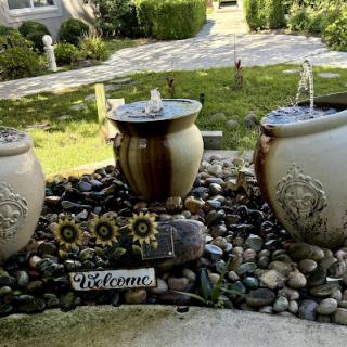 A serene garden setup featuring three decorative water fountains surrounded by smooth pebbles and a welcoming sign.