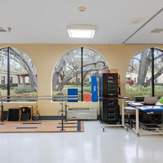 A spacious physical therapy room featuring exercise equipment and large windows that provide natural light.