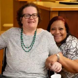 Two friends smiling together indoors, one seated in a wheelchair.