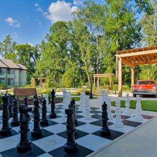 A scenic outdoor setting featuring a giant chessboard surrounded by trees, with a house and pergola in the background.