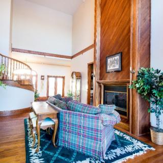 A spacious living room featuring a high ceiling, wooden accents, and a central fireplace, with a cozy seating area and staircase.