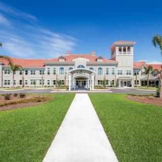 A grand building with red roof tiles and a central tower, surrounded by palm trees and a well-manicured lawn, under a clear blue sky.