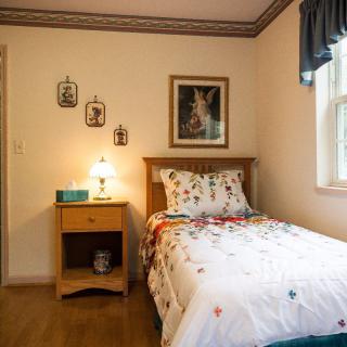 A cozy bedroom featuring a single bed with floral bedding and a nightstand with a lamp, adorned with wall art and natural light from a window.