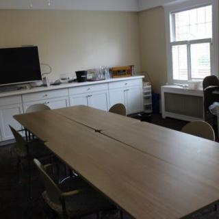 A meeting room featuring a long conference table with chairs, a television, and windows providing natural light.