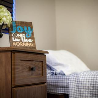 A cozy bedroom corner featuring a wooden nightstand with flowers and an inspirational sign, next to a neatly made bed.