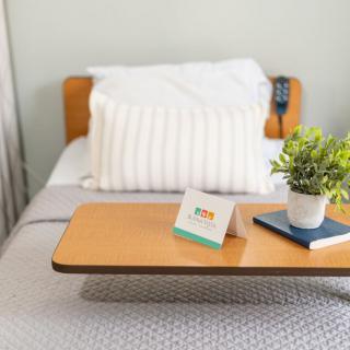 A cozy bed setup with a pillow, a tray table holding a plant and a card, alongside a book.