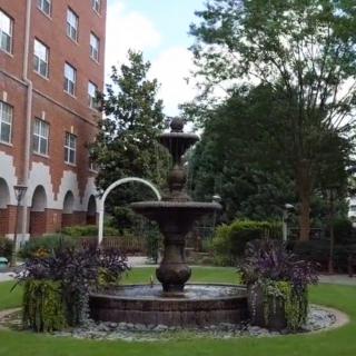 A serene garden featuring a beautiful fountain surrounded by lush greenery and a brick building.