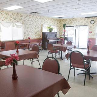 A spacious dining area featuring neatly arranged tables, chairs, and decorative flowers, with a piano in the background.