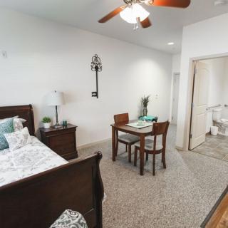 A cozy bedroom with a wooden bed, a small dining table with chairs, and a ceiling fan.