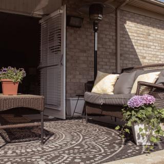 A cozy patio area with rattan furniture, potted flowers, and a patterned rug against a brick wall.