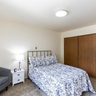 A cozy bedroom with a patterned bedspread, nightstand with lamp, chair, and wooden closet.