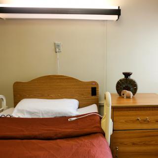 A simple hospital room featuring a wooden bed with white linens, a nightstand, and a decorative vase.