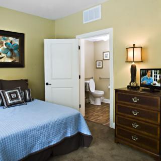 A cozy bedroom with a blue bedspread, wooden dresser, and an open door leading to a bathroom.