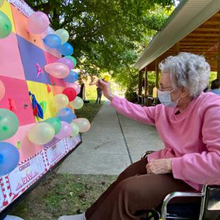 An elderly woman in a wheelchair enjoys an outdoor activity, interacting with colorful balloons.