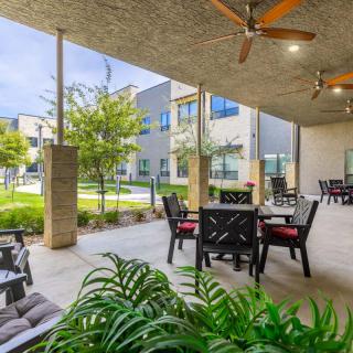 A modern outdoor patio with comfortable seating and ceiling fans, surrounded by greenery and contemporary buildings.