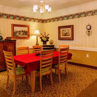 A cozy dining room featuring a wooden table with a red tablecloth, surrounded by six chairs, illuminated by a chandelier and accented with wall art and decorative wallpaper.