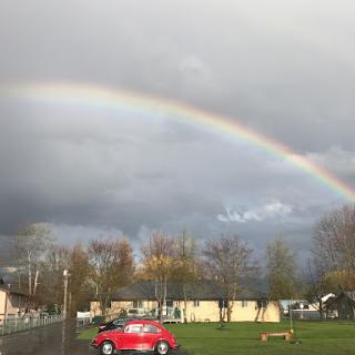 A vibrant rainbow arching over a serene landscape after a rain, with a red car parked below.