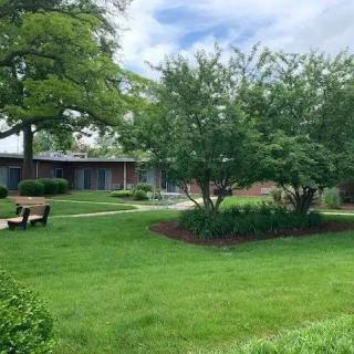 A tranquil park scene with a bench, trees, and a building in the background under a partly cloudy sky.