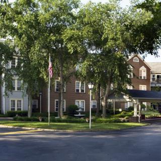 A large brick building surrounded by trees, featuring a driveway with a flagpole at the entrance.