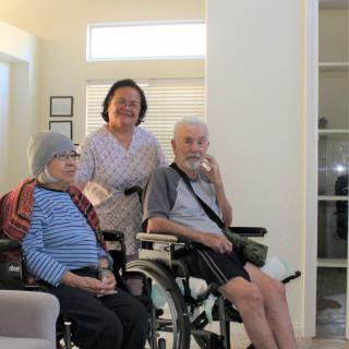 An elderly couple in wheelchairs, accompanied by a caregiver, in a welcoming indoor environment.