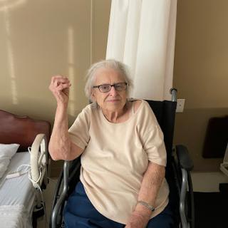 An elderly woman sits in a wheelchair, smiling warmly in her cozy room.