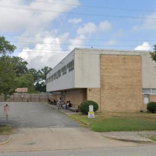 A view of a school building with a parking area and trees under a cloudy sky.