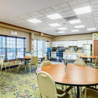 A spacious kitchen and dining area with multiple round tables and chairs, well-lit by overhead lights and natural light from large windows.