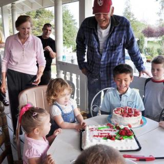 A group of children and adults gather around a table with cakes during a birthday celebration on a porch.