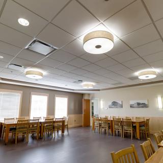 A well-lit dining room with several tables and chairs, featuring ceiling lights and large windows.
