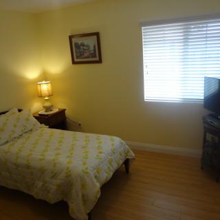 A cozy bedroom featuring a single bed, a bedside lamp, a window with blinds, and a television on a stand.