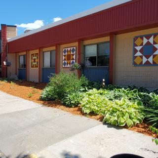 A sunny view of a building exterior decorated with quilt patterns and surrounded by landscaped greenery.