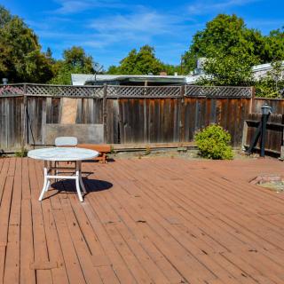 A sunny backyard patio with a wooden deck, featuring a table and chair set against a backdrop of trees and a fence.