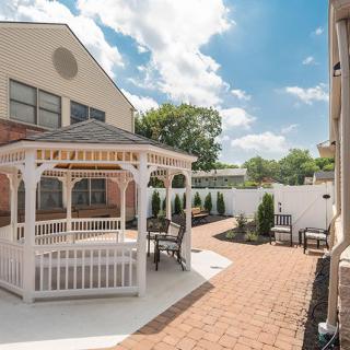 A serene residential backyard featuring a white gazebo, brick pathway, and comfortable outdoor seating under a partly cloudy sky.