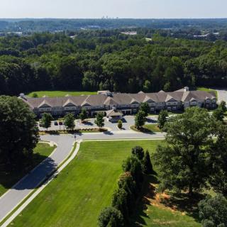 Aerial view of a building surrounded by greenery and trees, with a visible roadway and parking lot.