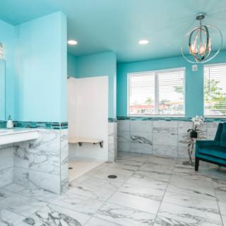 A modern bathroom with marble flooring and turquoise walls, featuring a luxurious armchair and chandelier.