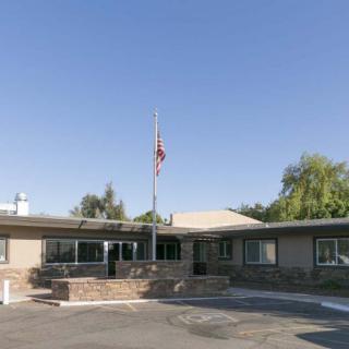 A one-story office building with an American flag in front, surrounded by a parking lot and trees under a clear blue sky.