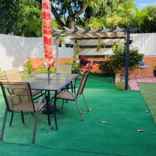 A cozy backyard patio featuring a table with chairs under a colorful umbrella, adjacent to a pergola surrounded by lush greenery.