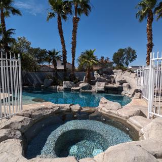 A scenic outdoor area featuring a pool and hot tub surrounded by palm trees and rocks, enclosed by white fencing.